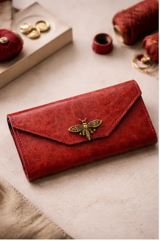 Lähikuva Toe-Nahka Designin punaisesta, tekstuuripintaisesta nahkalompakosta, jossa on koristeellinen kullanvärinen mehiläissolki. Lompakko on asetettu työpöydälle, jonka taustalla näkyy lankarullia ja käsityövälineitä.